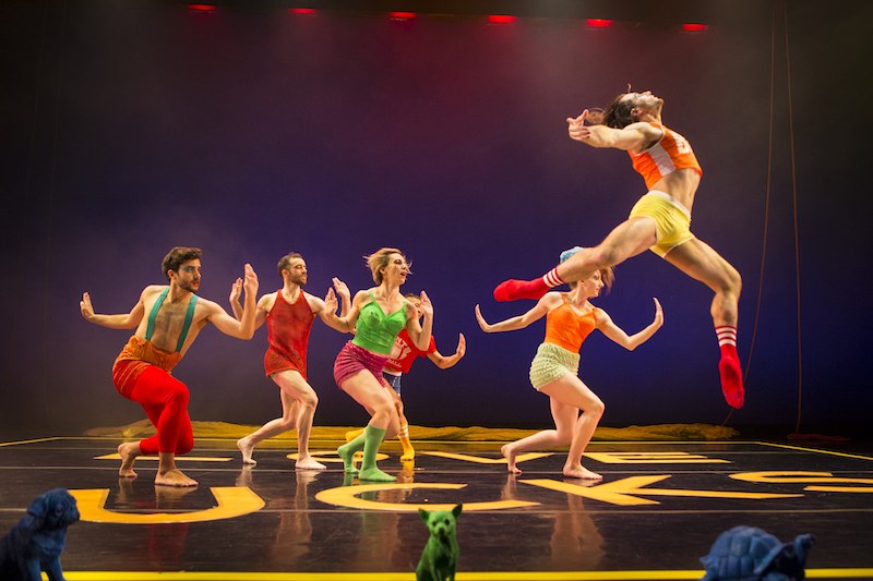 A dancer in the foreground leaps high into the air; his arms outstretched and head and chest lift to the ceiling. A group of dancers in the background assume a lunge position with one arm flexed at the elbow in front of their body and the other bent behind them.